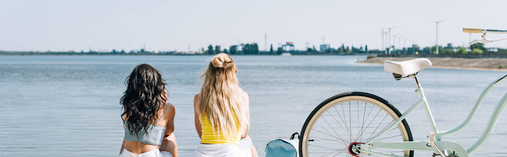 Widok z tyłu Blondynka i brunetka przyjaciele siedzący w pobliżu rower i rzeka w lato, panoramiczny strzał - Zdjęcie, obraz