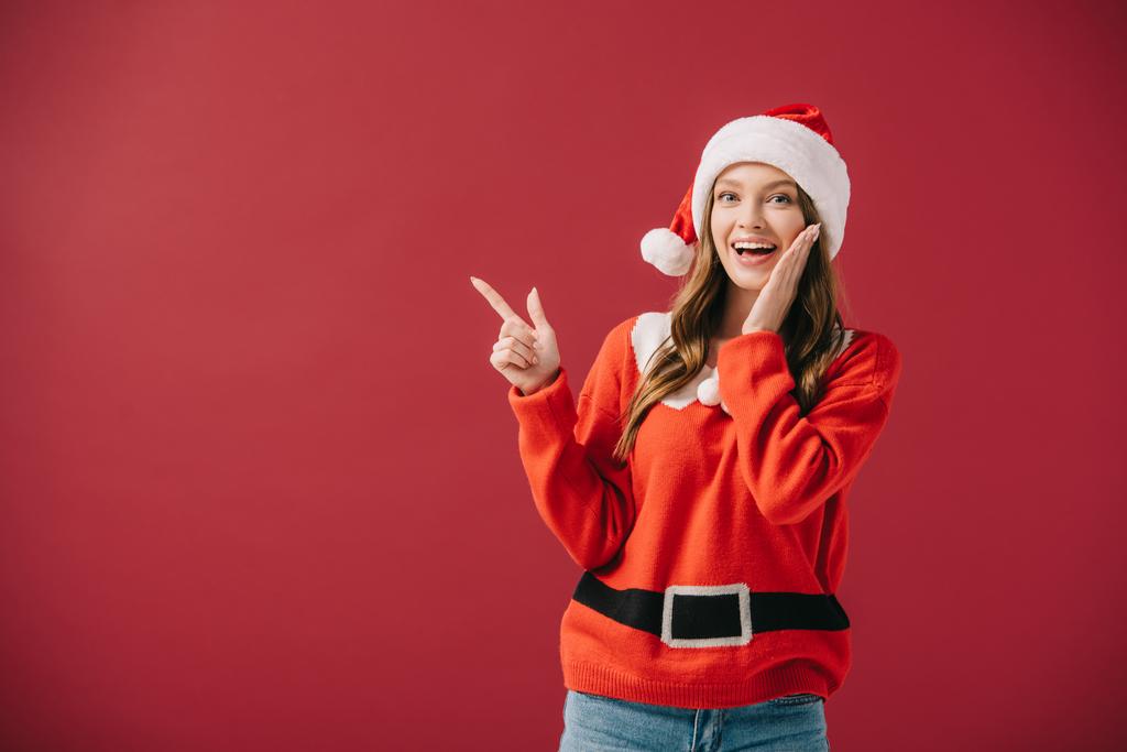 vonzó nő Santa kalap és pulóver mutatva ujjal izolált piros  - Fotó, kép