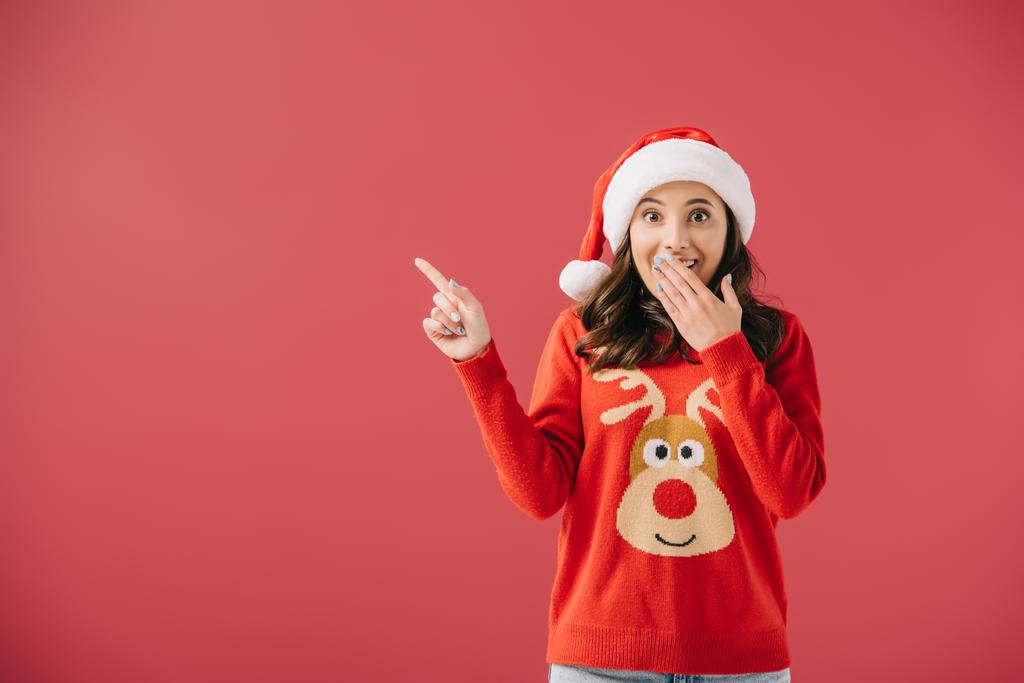 attraktive und schockierte Frau mit Weihnachtsmütze und Pullover, die mit dem Finger auf rot zeigt  - Foto, Bild
