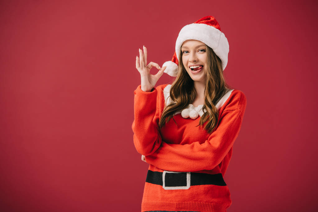 attraktive Frau mit Weihnachtsmütze und Pullover, die auf rotem Grund vereinzelt OK-Geste zeigt  - Foto, Bild