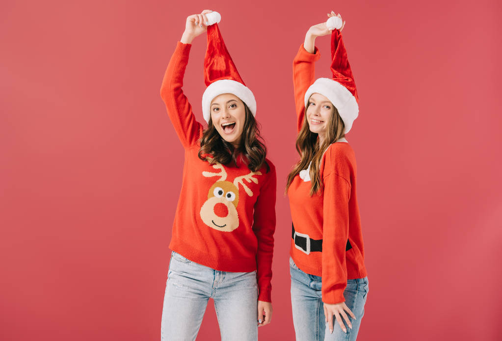 aantrekkelijke en lachende vrouwen in truien houden van Santa hoeden geïsoleerd op rood  - Foto, afbeelding