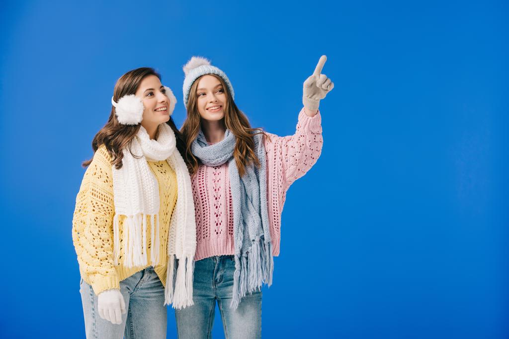 привлекательные и улыбающиеся женщины в свитерах и шарфах, указывающие пальцем и смотрящие в сторону, изолированные на голубом
 - Фото, изображение