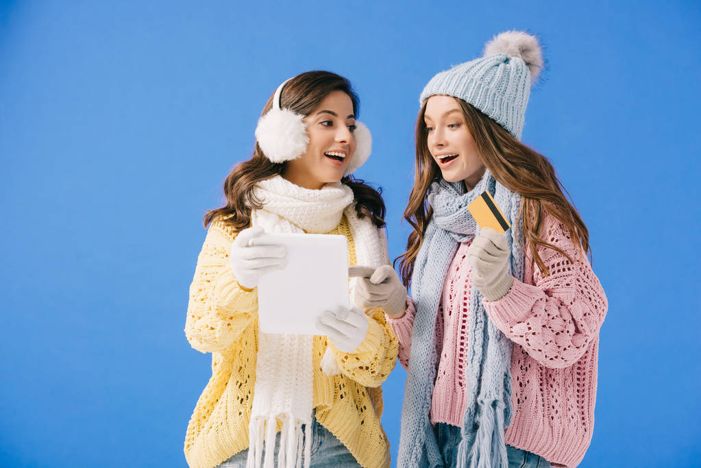 attractive and smiling women in sweaters and scarves holding digital tablet and looking at camera isolated on blue - Photo, Image