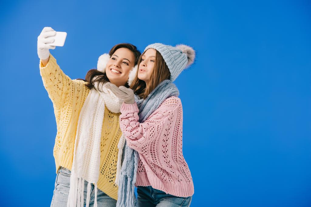 kazak ve eşarp çekici ve gülümseyen kadınlar selfie alarak ve mavi izole öpücük üfleme - Fotoğraf, Görsel