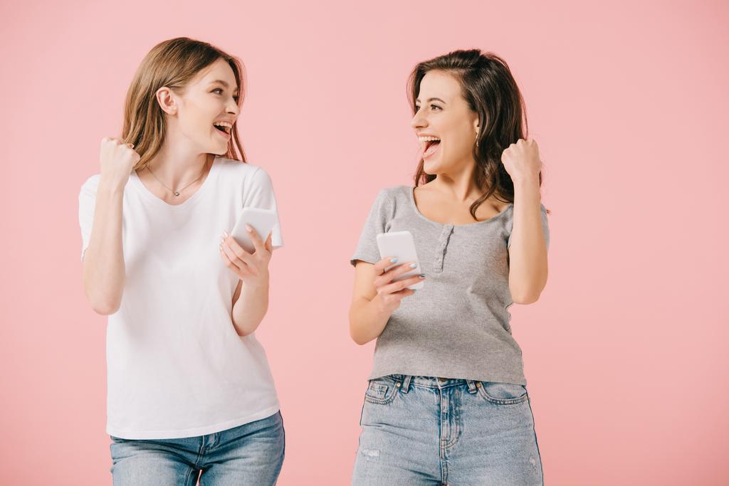 ελκυστικές και χαμογελαστές γυναίκες σε μπλουζάκια που κρατούν smartphones και δείχνουν Ναι χειρονομία απομονωθεί σε ροζ - Φωτογραφία, εικόνα