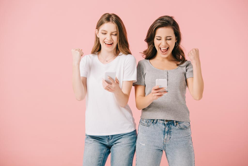 ελκυστικές και χαμογελαστές γυναίκες σε μπλουζάκια που κρατούν smartphones και δείχνουν Ναι χειρονομία απομονωθεί σε ροζ - Φωτογραφία, εικόνα