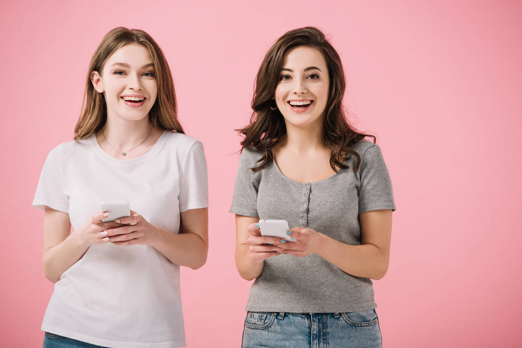 привабливі і усміхнені жінки в футболках тримають смартфон ізольовані на рожевому
 - Фото, зображення
