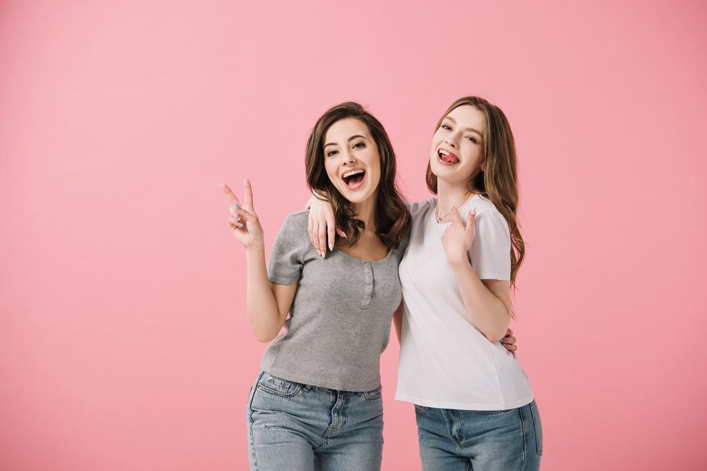 atraktivní a usmívající se ženy v tričkách, které se dívají na kameru a ukazují mírovou známku izolovanou na růžovém - Fotografie, Obrázek