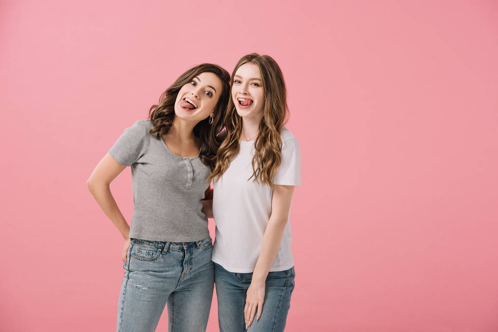 ελκυστικές και χαμογελαστές γυναίκες σε μπλουζάκια με το να προεξέχουν γλώσσες κοιτάζοντας την κάμερα απομονωμένη σε ροζ - Φωτογραφία, εικόνα
