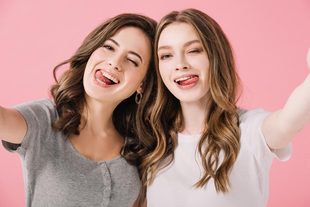 attraktive und lächelnde Frauen in T-Shirts mit herausgestreckten Zungen, die vereinzelt in die Kamera auf rosa sehen - Foto, Bild