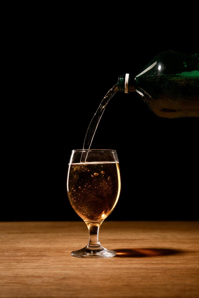 beer pouring from bottle in glass on wooden table isolated on black - Photo, Image