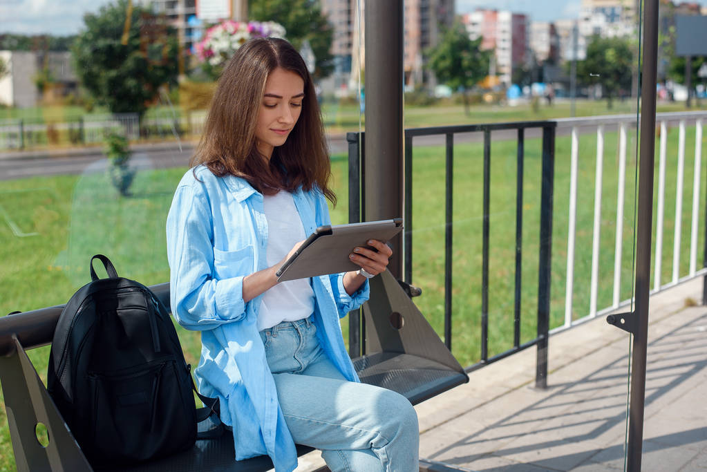 glückliche Frau benutzt Tablet oder E-Book an einer Straßenbahnhaltestelle, während sie auf öffentliche Verkehrsmittel wartet. - Foto, Bild