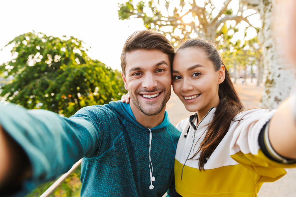 Foto eines lächelnden jungen Mannes und einer lächelnden Frau, die Selfie-Fotos machen und sich umarmen, während sie auf dem Stadtboulevard trainieren - Foto, Bild