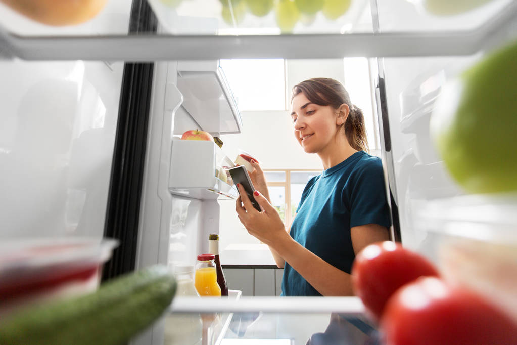 Frau mit Smartphone macht Liste mit Lebensmitteln im Kühlschrank - Foto, Bild