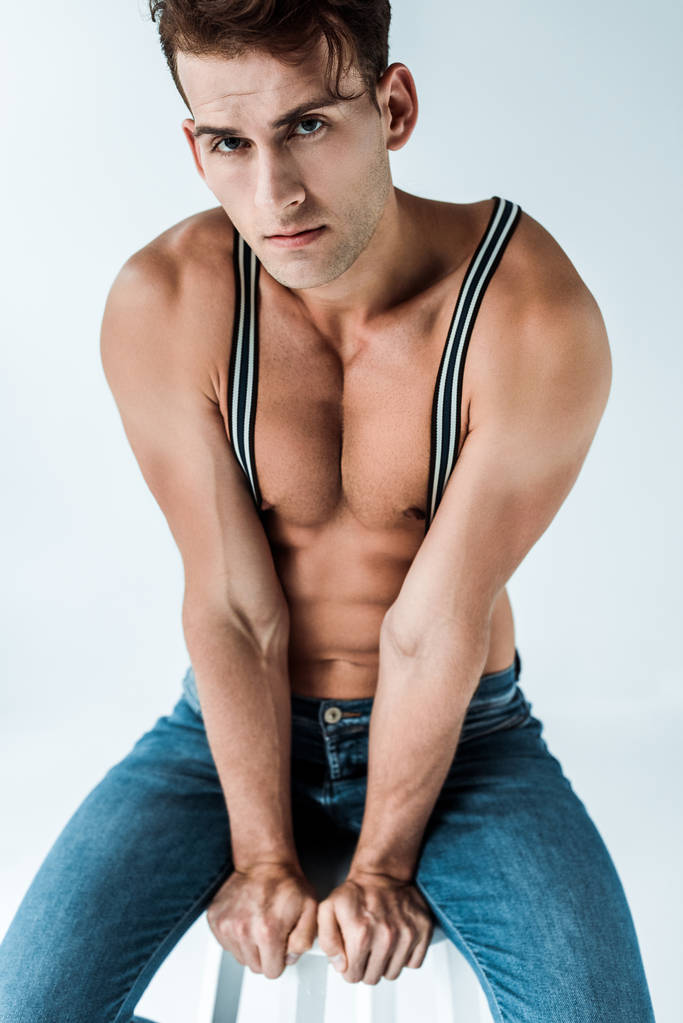 sexy man in suspenders sitting on chair and looking at camera on white  - Photo, Image