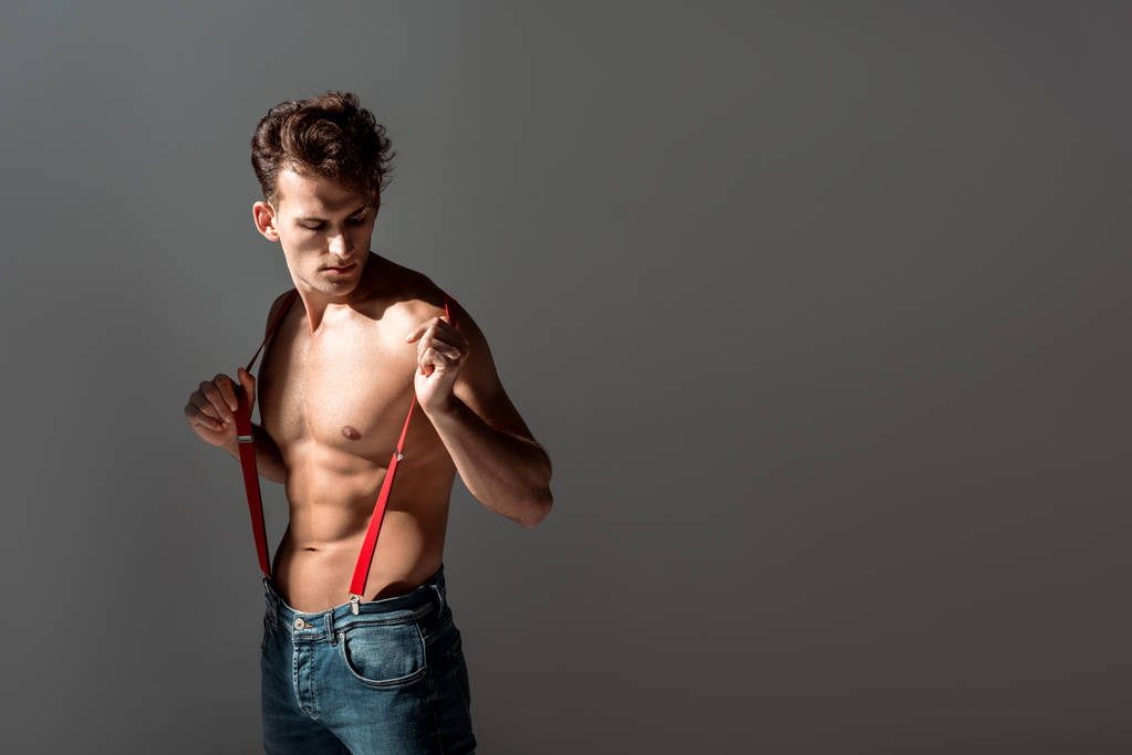 shirtless and handsome man looking at red suspenders while standing isolated on grey   - Photo, Image