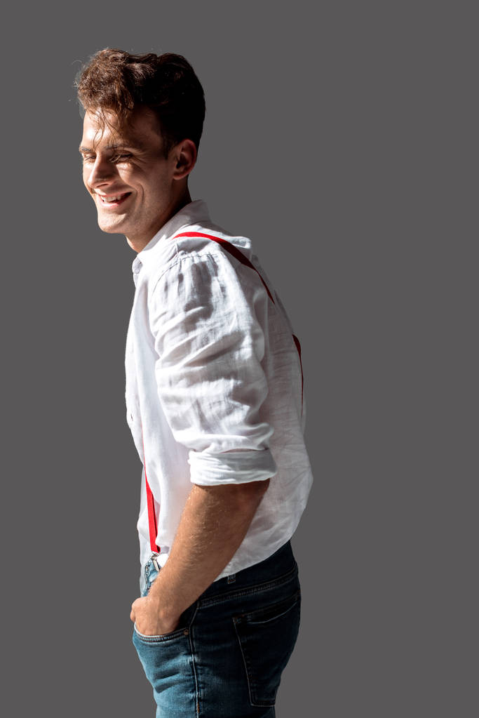 szczęśliwy młody człowiek w białej koszulce stojący z rękami w kieszeniach wyizolowanych na szaro  - Zdjęcie, obraz
