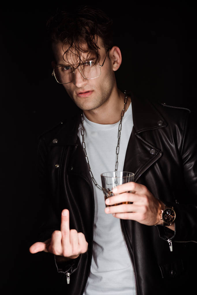selective focus of stylish man in eye glasses holding whiskey and showing middle finger isolated on black  - Photo, Image