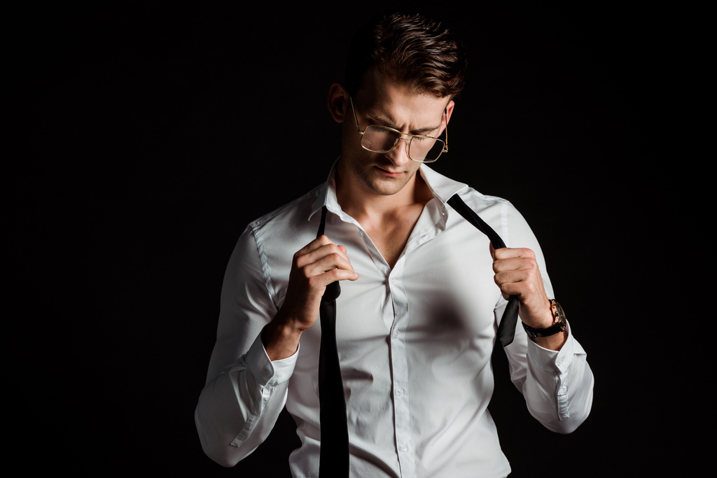 pohledný obchodník s brýlemi, který bere kravatu izolovaný na černém  - Fotografie, Obrázek