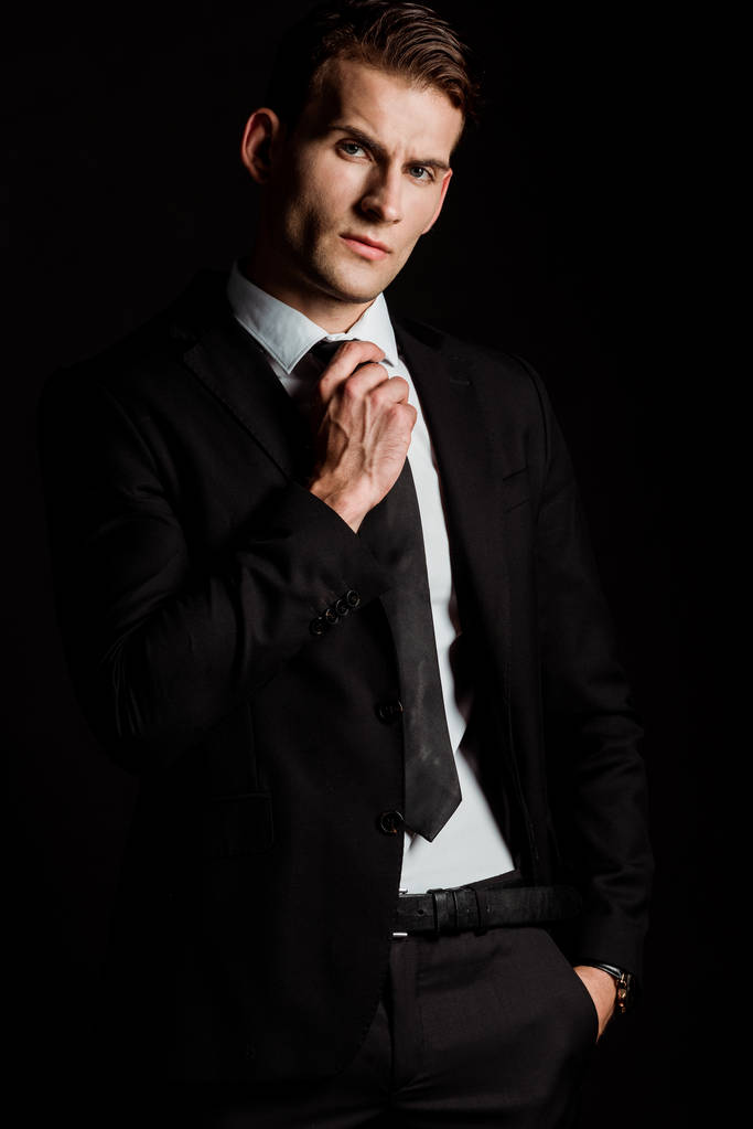 красивий бізнесмен торкається краватки, стоячи з рукою в кишені ізольовані на чорному
  - Фото, зображення