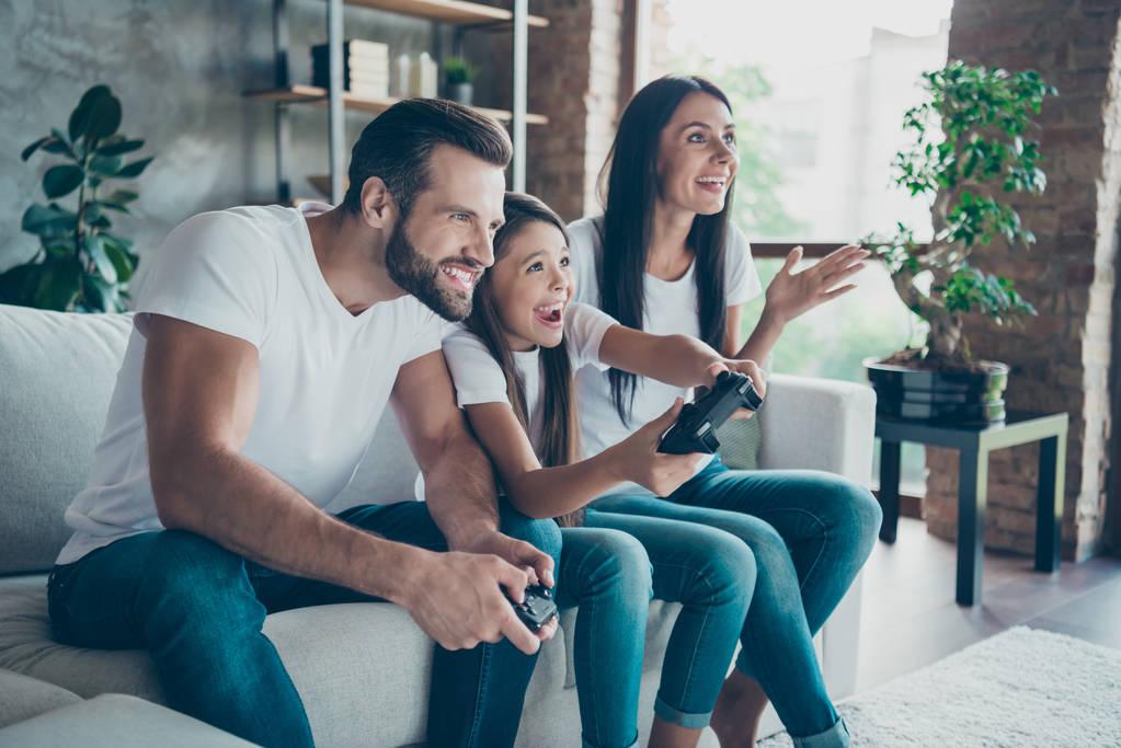 Профиль вид сбоку красивой привлекательной веселой веселой семьи носить случайные белые футболки джинсы джинсы сидя на диване с веселыми игроками наркоманов с использованием геймпада в помещении
 - Фото, изображение