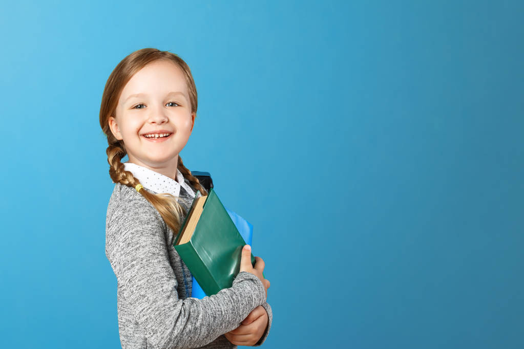 Крупный план портрета маленькой девочки школьницы на синем фоне. Ребенок держит книги. Концепция образования и школы. Копирование пространства
 - Фото, изображение