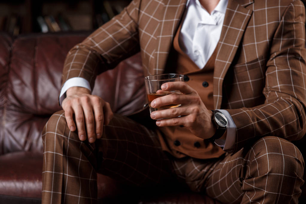 Młody, odnoszący sukcesy mężczyzna w brązowym kosmtyum pije whisky. brodaty biznesmen siedzący w bibliotece na luksusowej skórzanej kanapie i pijący koniak - Zdjęcie, obraz