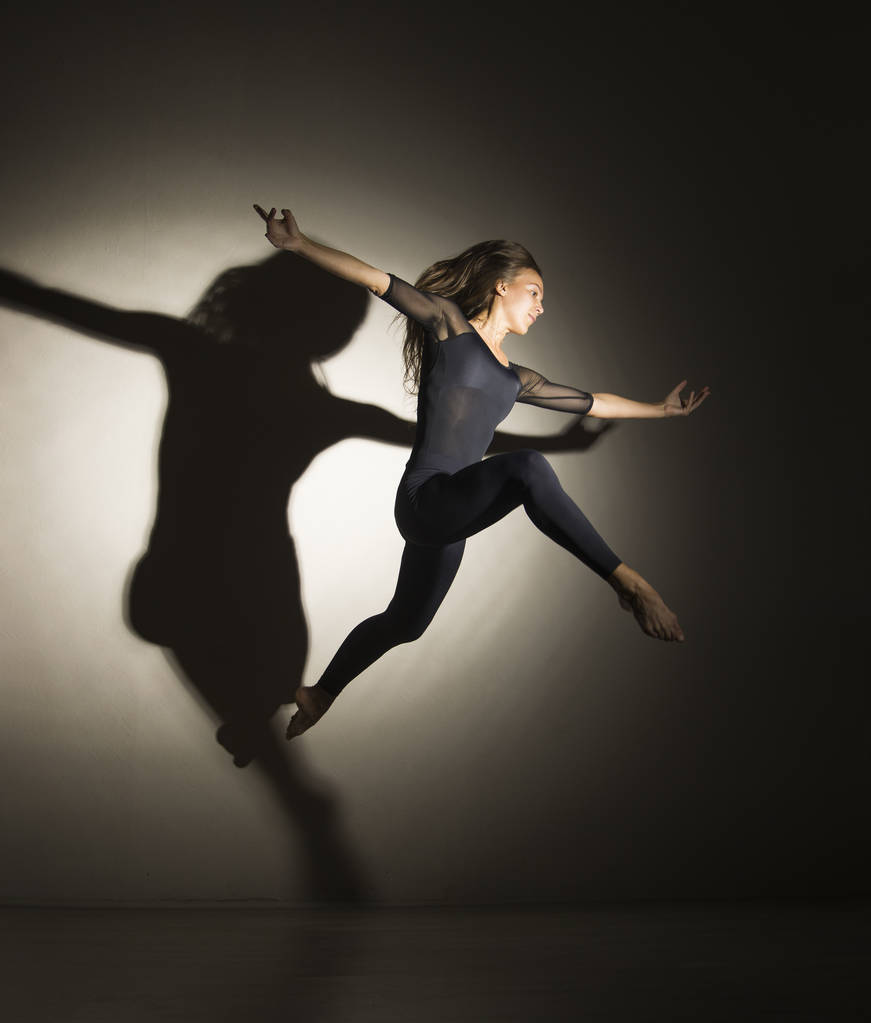 Дівчина в темряві виконує гімнастичні стрибки, на білому тлі є тінь з форми
. - Фото, зображення