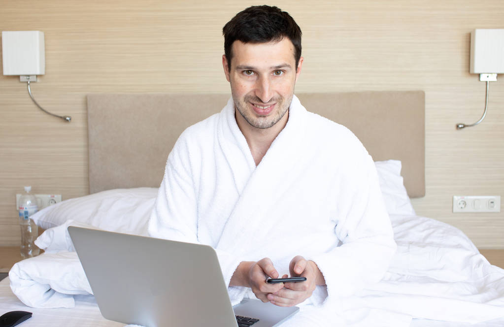 Jonge man in badjas met laptop en smartphone in de slaapkamer 's ochtends - Foto, afbeelding