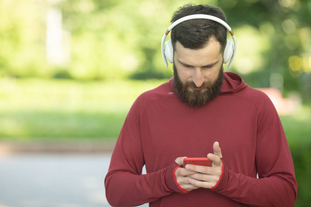 Bearded man in headphones and sportswear using smartphone during training in park - Photo, Image