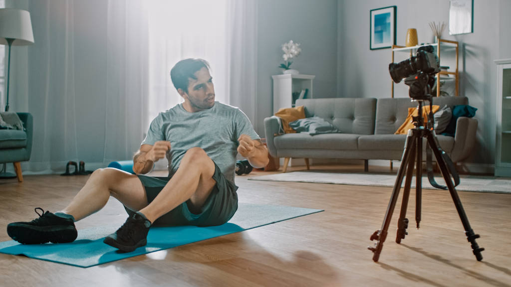 Ein kräftiger, athletischer Mann in T-Shirt und Shorts hält sein Workout für seinen Blog mit der Kamera fest. Szene spielt in seinem geräumigen und hellen Wohnzimmer mit minimalistischem Interieur. - Foto, Bild