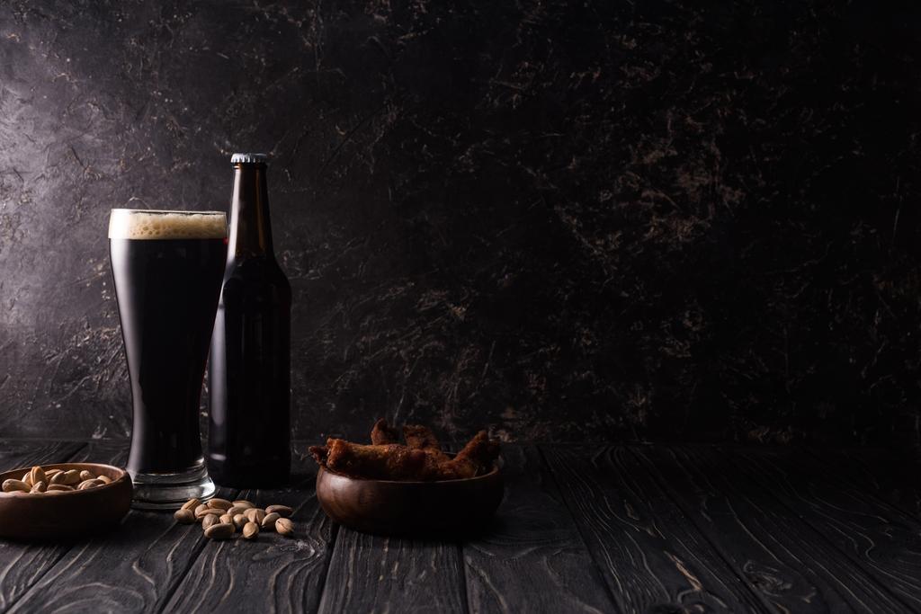 стекло и бутылка тёмного пива возле миски с фисташками и куриными крылышками на деревянном столе
 - Фото, изображение