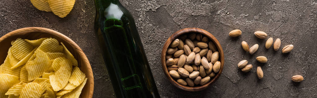 panoramic shot of bottle of beer near bowls with pistachios and crisps on brown textured surface - Photo, Image