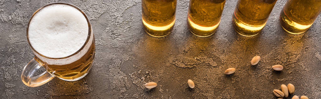 tiro panorâmico de garrafas de cerveja leve perto de pistácios espalhados na superfície texturizada marrom
 - Foto, Imagem