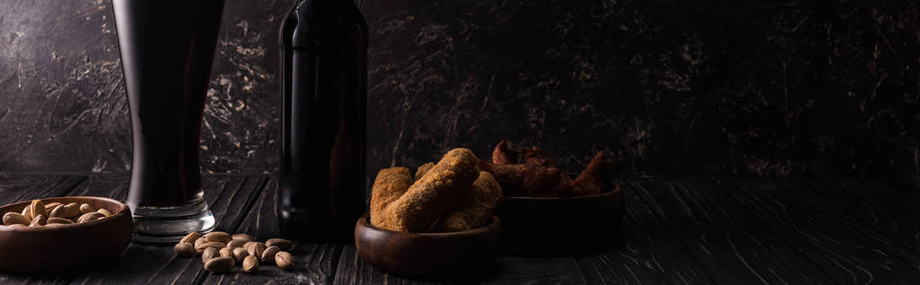 tiro panorâmico de garrafa e copo de cerveja escura perto de tigelas com lanches na mesa de madeira
 - Foto, Imagem