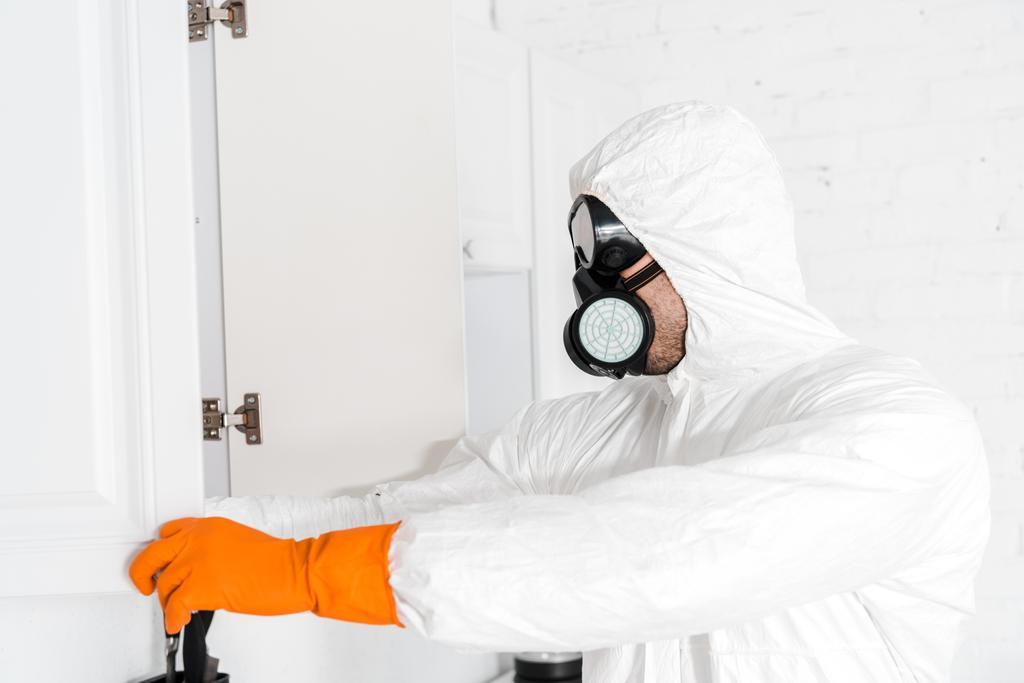 exterminator in protective mask and uniform standing near kitchen cabinet  - Photo, Image