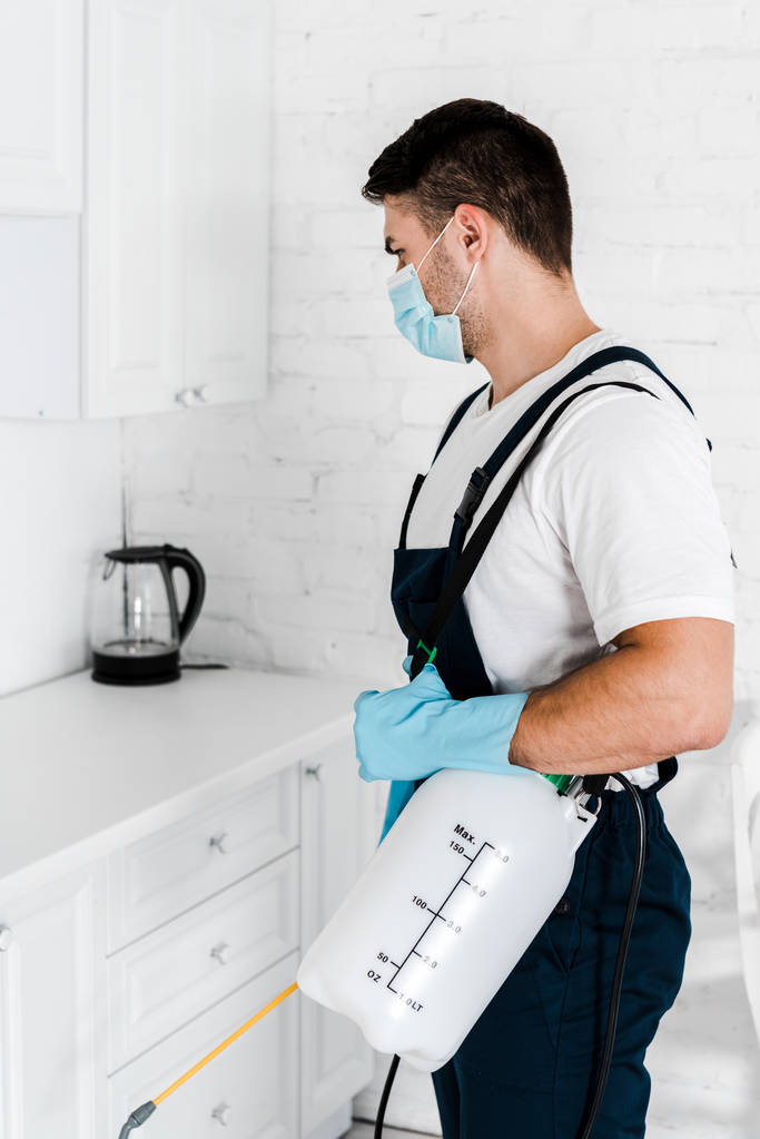 exterminator in protective mask and uniform holding toxic spray standing in kitchen  - Photo, Image