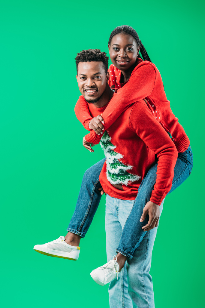 Αφρικανός Αμερικανός άνθρωπος κρατώντας στην πλάτη γυναίκα σε χριστουγεννιάτικο πουλόβερ απομονωθεί σε πράσινο - Φωτογραφία, εικόνα