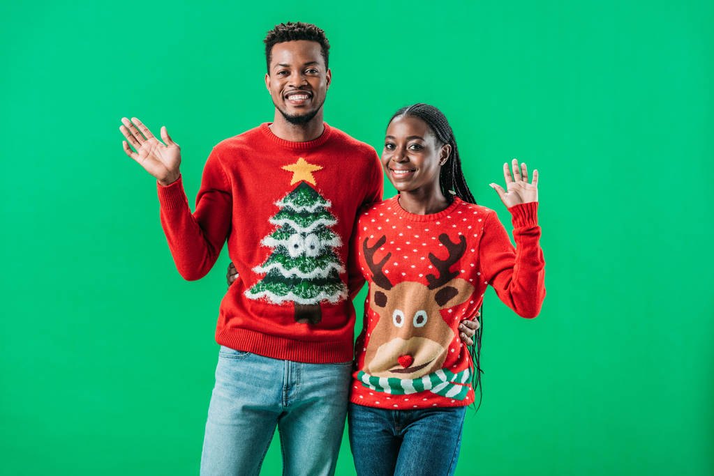 Афроамериканец мужчина и женщина в рождественских свитерах машут руками и смотрят на камеру, изолированную на зеленом
 - Фото, изображение