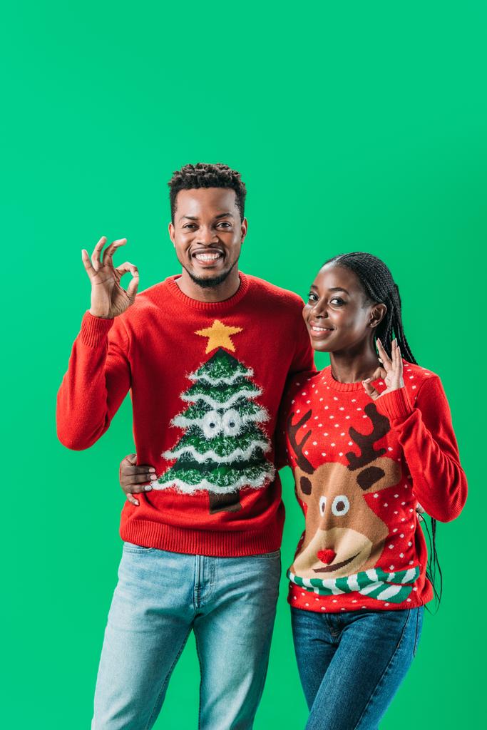 Africano americano homem e mulher em camisolas de Natal mostrando sinal ok e olhando para a câmera isolada no verde
 - Foto, Imagem