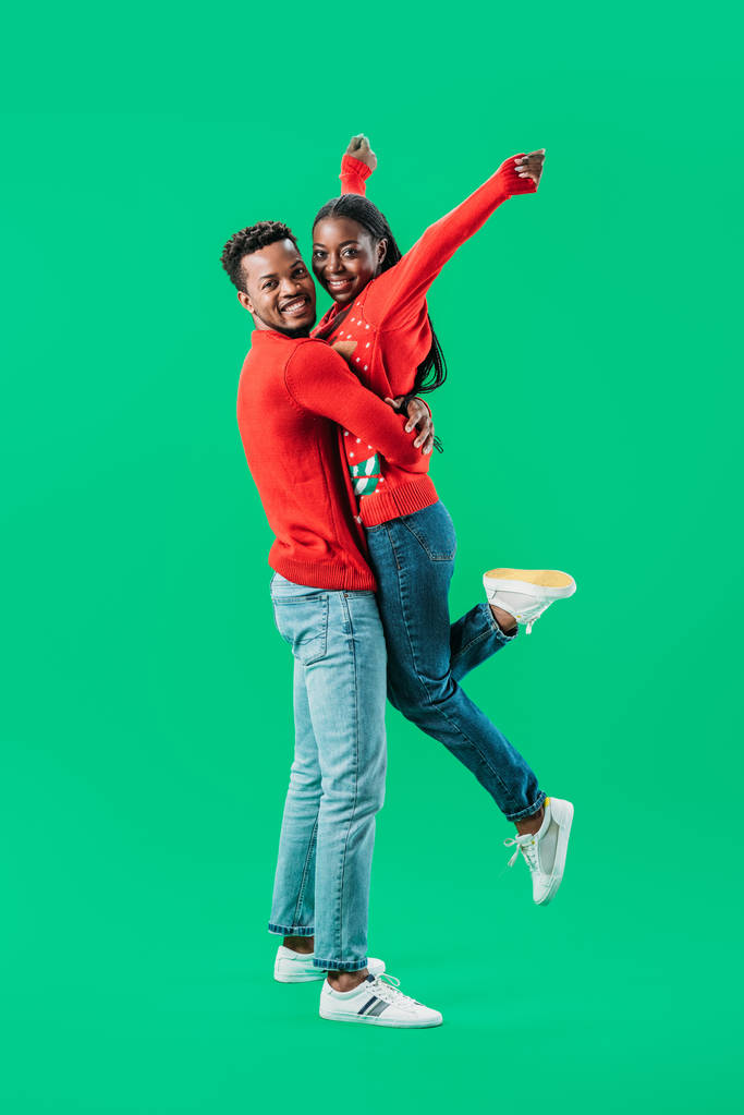 赤いクリスマスセーターを着たアフリカ系アメリカ人男性が、緑色に孤立した手を挙げた女性を抱きしめる - 写真・画像