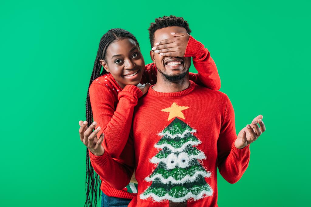 African American woman in Christmas sweater covering eyes with hand to man isolated on green - Photo, Image
