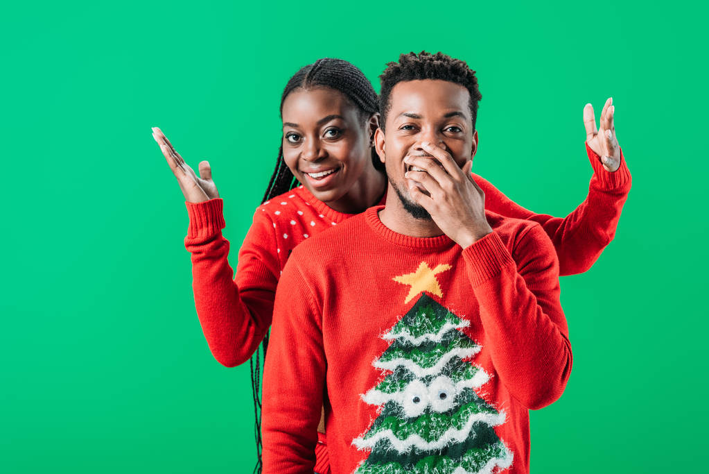 アフリカ系アメリカ人女性は、緑の上に孤立したクリスマスセーターで驚いた男の後ろに手を上げる - 写真・画像
