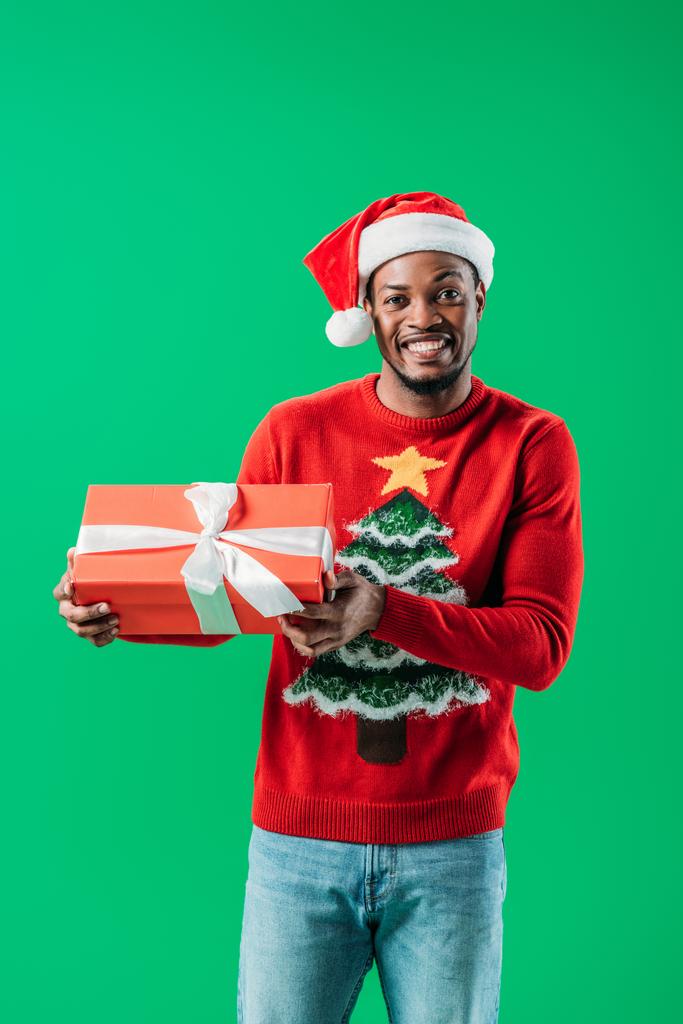 African American man in Christmas sweater and Santa hat holding red gift box looking at camera isolated on green - Photo, Image