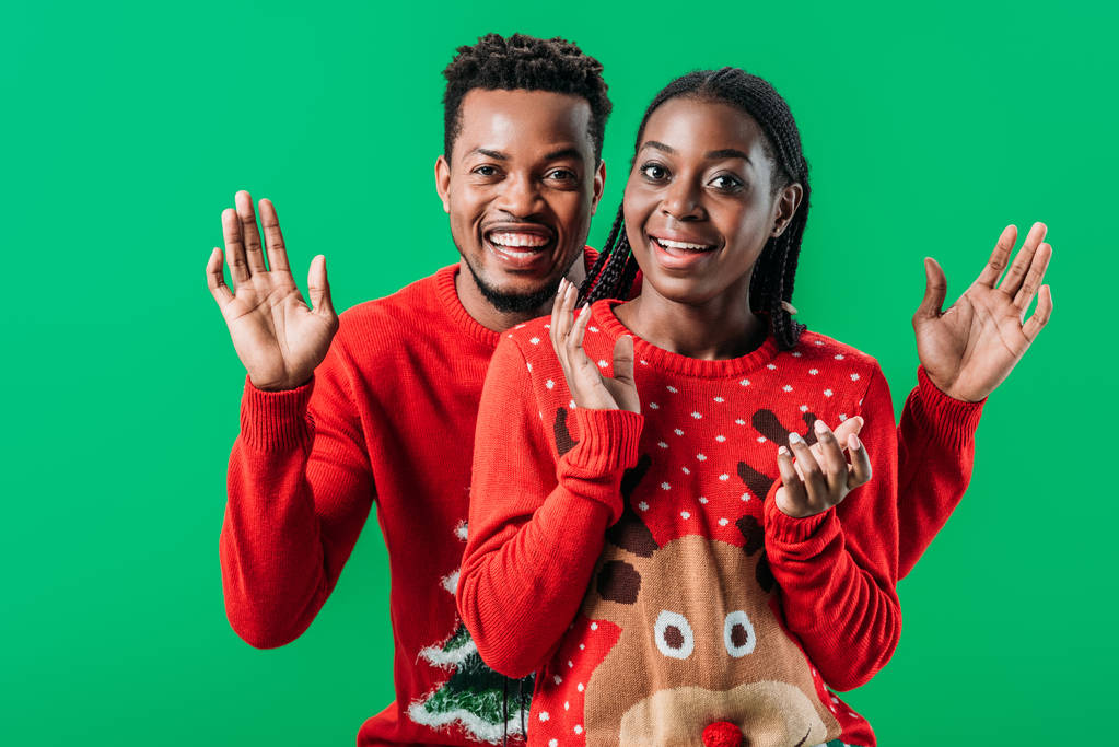 χαρούμενο αφρικανικό ζευγάρι με κόκκινα Χριστουγεννιάτικα πουλόβερ χαμογελώντας και κοιτώντας την κάμερα απομονωμένη στο πράσινο - Φωτογραφία, εικόνα
