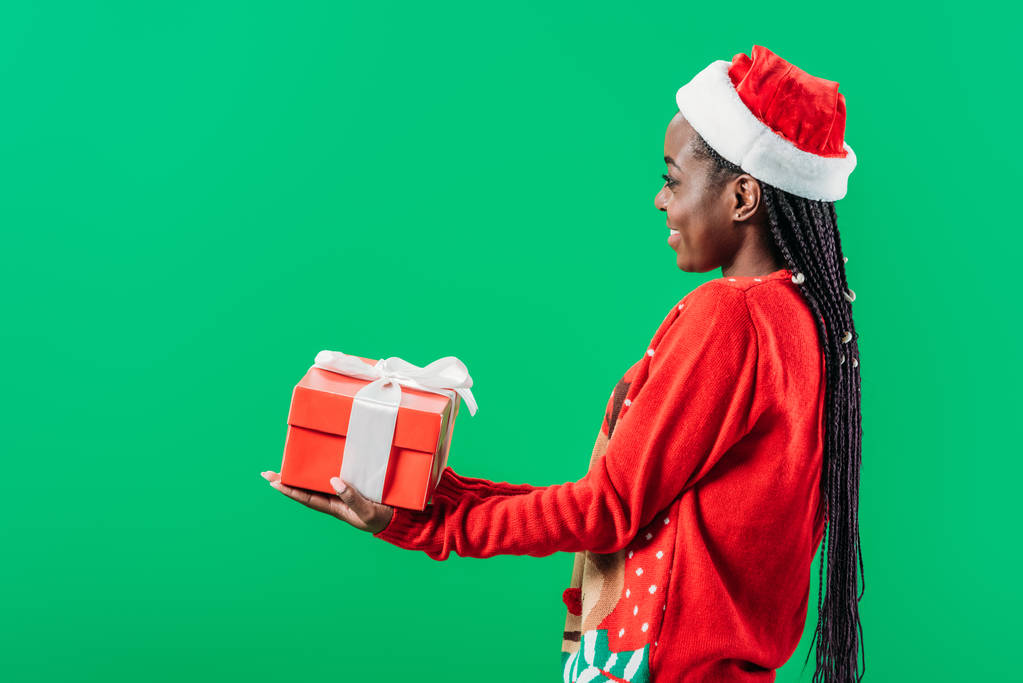side view of African American woman in Christmas sweater and Santa hat raising hands with gift box isolated on green - Photo, Image