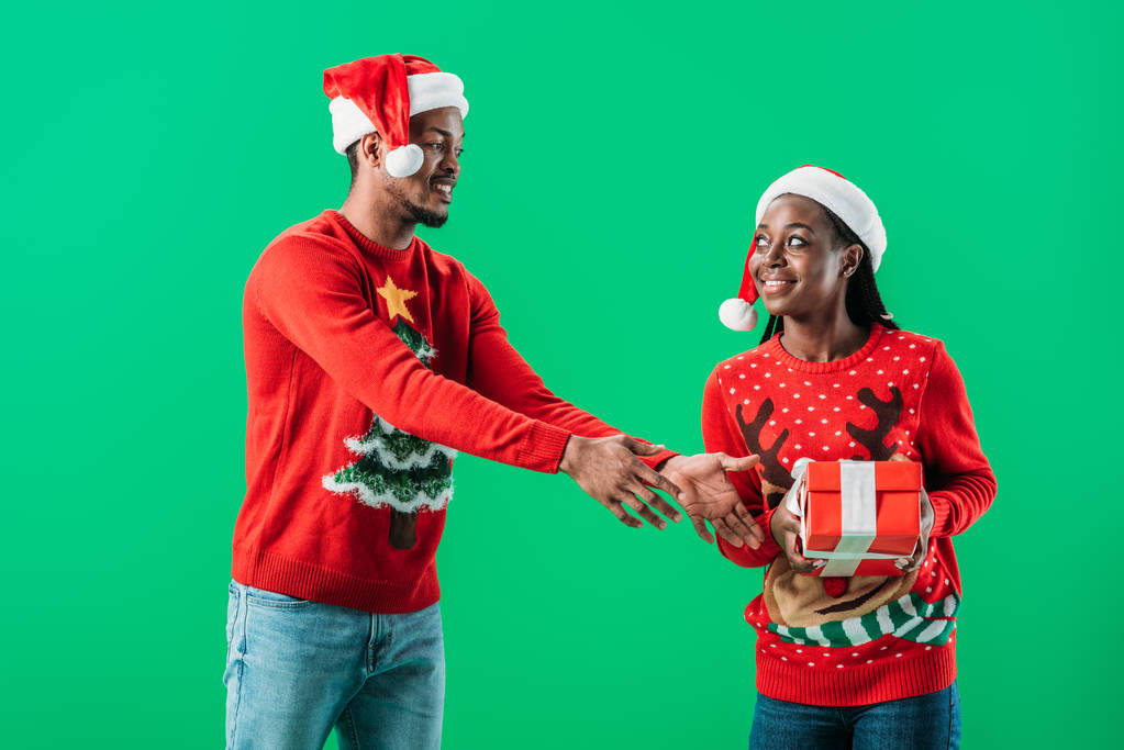 African American man in red Christmas sweater raising hands to gift box in hands of woman isolated on green - Photo, Image