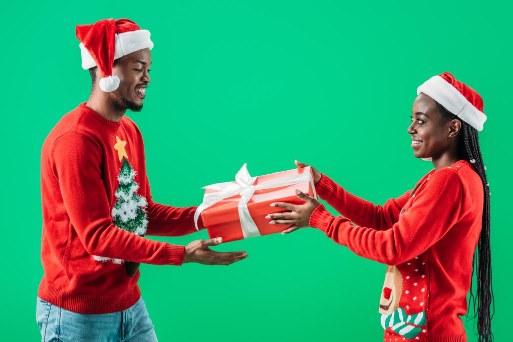 side view of African American man in red Christmas sweater getting gift box from woman isolated on green - Photo, Image