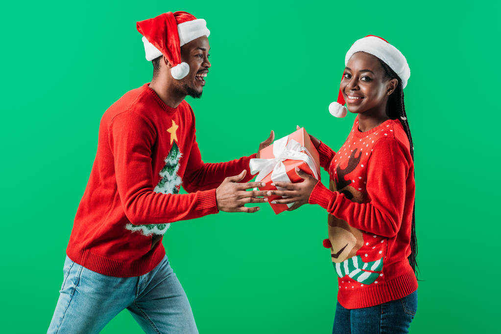 赤いクリスマスセーターとサンタ帽子のアフリカ系アメリカ人男性の側面図は、緑の上に孤立した女性からギフトボックスを取得します - 写真・画像