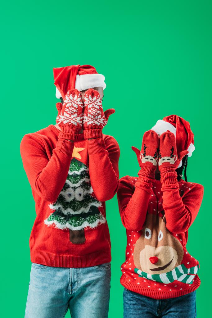Casal afro-americano em blusas vermelhas de Natal e chapéus de Papai Noel cobrindo os olhos com as mãos em luvas de inverno com padrão isolado no verde
 - Foto, Imagem
